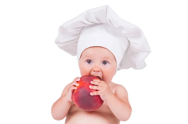 Uma criança pequena está comendo um grande pêssego em um terno de chef em um fundo branco . — Fotografia de Stock