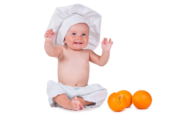 Un bambino piccolo mangia una fetta d'arancia in un abito da chef su uno sfondo bianco . — Foto Stock