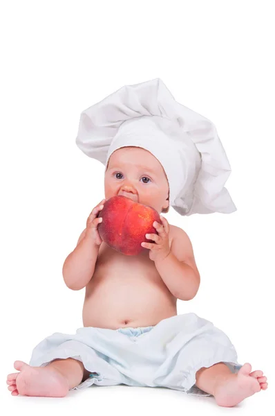 Ein kleines Kind isst einen großen Pfirsich im Kochanzug auf weißem Hintergrund. — Stockfoto