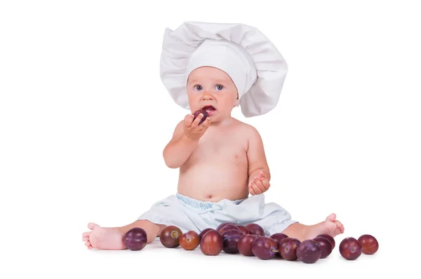 Un bambino piccolo sta mangiando prugne in un abito da chef su uno sfondo bianco . — Foto Stock