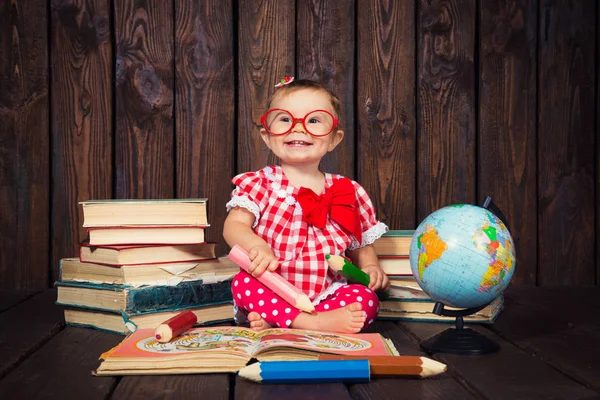 Счастливая маленькая девочка с очками и ручкой на фоне книг и глобуса — стоковое фото
