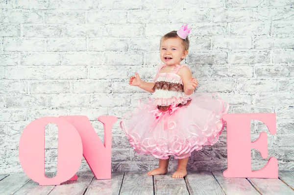 Schöne und süße Mädchen in rosa Kleid auf dem Hintergrund des Wortes eins, weint an ihrem ersten Geburtstag. emotionales Kind. — Stockfoto