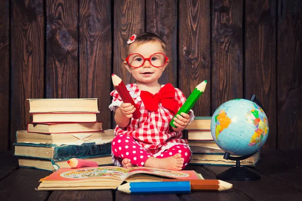 Happy a nice little girl with glasses and pencils against the background of books and a globe — Stock Photo, Image