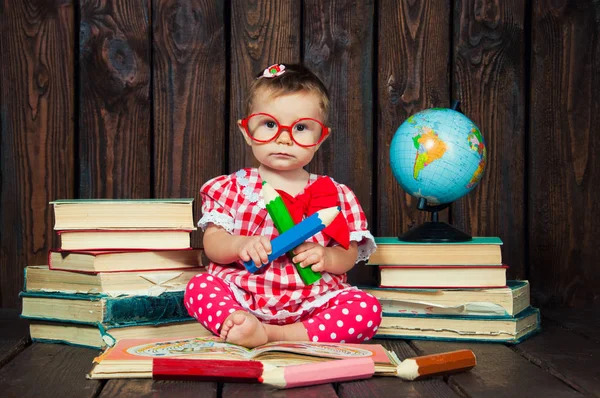 Happy a nice little girl with glasses and pencils against the background of books and a globe — Stock Photo, Image