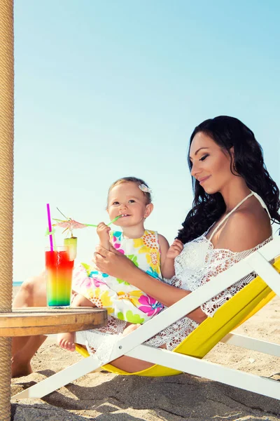 Junge schöne Mutter und ihre kleine Tochter am Strand und haben Spaß. die Mädchen trinken einen Cocktail — Stockfoto