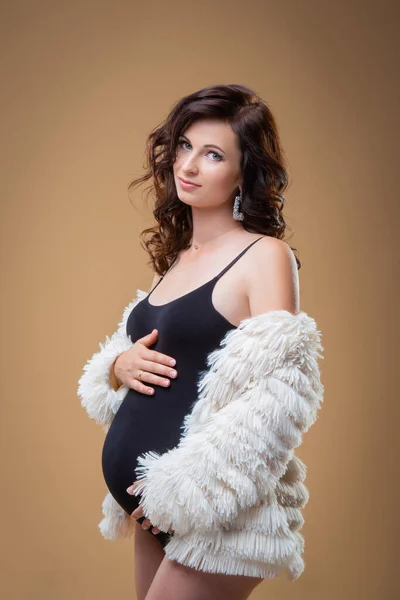 Jonge brunette zwangere vrouw in bontjas op een effen achtergrond. Mooie stijlvolle gezonde zwangerschap. Modekleding — Stockfoto
