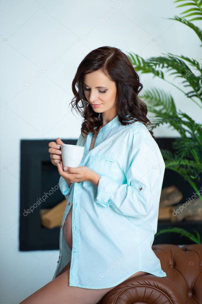 Beautiful pregnant brunette woman in a shirt sits on a sofa in a living room, holds a cup and drinks tea