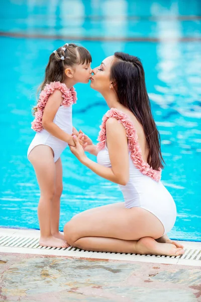 Havuzun kenarında oturan aynı beyaz mayo giymiş küçük kızıyla genç bir anne. Sevgiler ve öpücükler Yaz ve deniz aileleri için kıyafetler. — Stok fotoğraf
