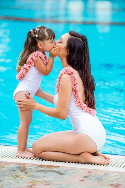 Giovane mamma con bambina con gli stessi costumi da bagno bianchi seduti sul bordo della piscina. Amore e bacio. Abbigliamento per estate e mare family look — Foto Stock