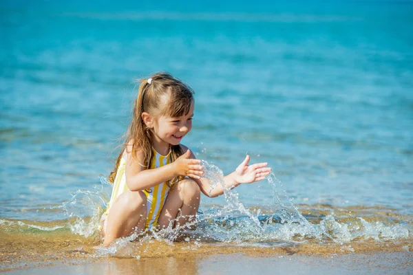 3 yaşında mayo giymiş bir kız kumlu deniz kıyısında oturur ve denizle oynar, su sıçratır. — Stok fotoğraf