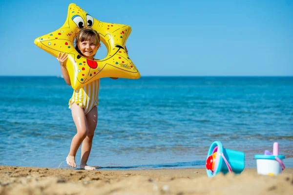 Słodka dziewczynka w stroju kąpielowym z nadmuchiwaną rozgwiazdy w dłoniach grimaces i uśmiecha się szczęśliwie na plaży morskiej. Pozytywny nastrój, szczęśliwe wakacje i zabawa — Zdjęcie stockowe