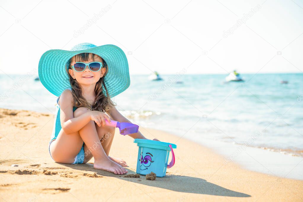 Little child girl fashionista in a blue wide-brimmed hat and sunglasses sits on the sand with a bucket on the seashore.