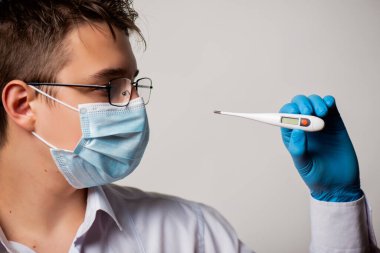 A doctor in a glasses and surgical mask holds an electronic thermometer to measure temperature. Signs of flu COVID 19 Coronavirus. clipart