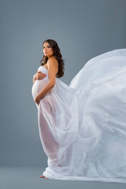 Stüdyo portresi, gri arka planda beyaz uçan kumaşlı hamile bir kadın. Güzel sağlıklı hamilelik, mutlu annelik..