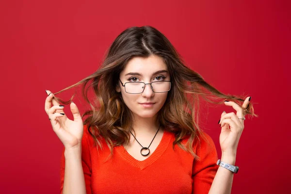 The girl in glasses twists hair with fingers on a red background.