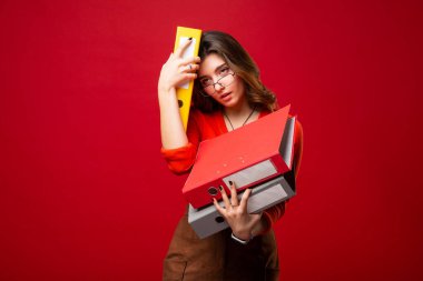 Gözlüklü kız kırmızı arka planda paniğe kapılmış klasörler taşıyor. Ruh hali stresi, muhasebe raporu, vergi denetimi, sınavlar.