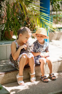 Küçük çocuklar evin merdivenlerinde oturur ve bir kediye, çocuklara ve hayvanlara olan sevgilerine sarılırlar.
