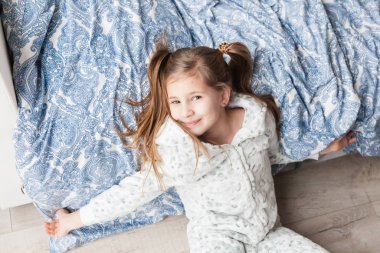 Açık gri pijamalı bir kız pencerenin yanındaki yatağa uzanmış, çocuk sabah uyanmış.