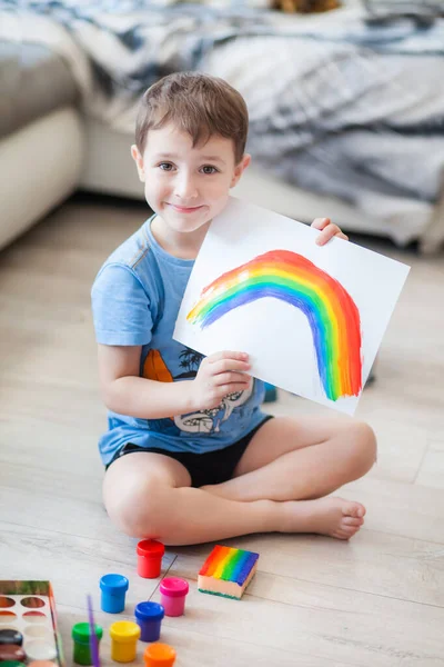 little boy draws a rainbow on a sheet of paper in different colors of gouache
