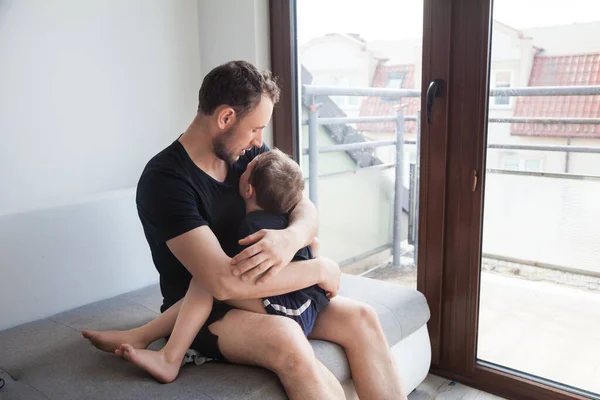 Baba oğluyla büyük bir pencerenin yanındaki gri bir kanepede, parlak bir odada oynuyor.