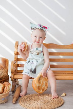 Bir yaşına kadar mutfak önlüğü takmış bir kız ve kafasında bir fiyonk bir bankta, rulolu turtaların yanında oturuyor, küçük bir fırıncı rulo ekmek yiyor.