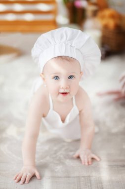 Bir yaşından küçük bir çocuk beyaz bir aşçı önlüğü ve şapkasıyla sepeti, ekmeği, çöreği, sütü ve unu olan bir bankta oturur.