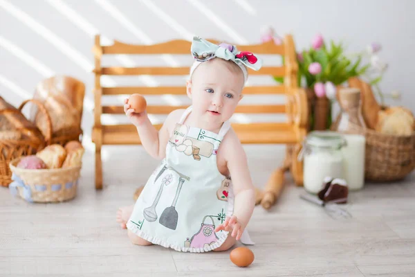 a little girl in a kitchen apron and a bow on her head sits near a bench, next to a basket with buns, bread and donuts, a bottle of milk and flour, a little baker eats buns