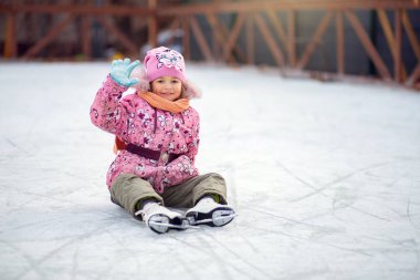 Artistik patenli bir kız buz pateni pistinde oturur ve elini sallar.