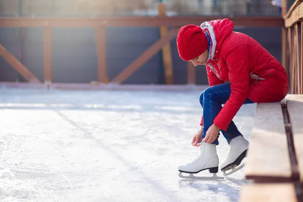 Chica en una chaqueta roja se sienta en un banco en una pista de hielo abierta y patines de encaje figura — Foto de Stock