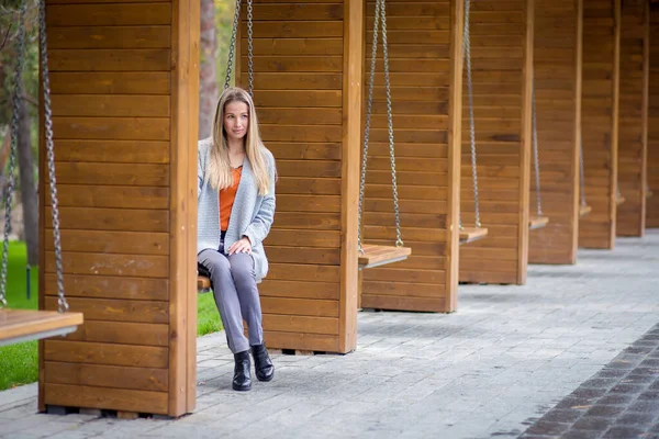 Schönes Mädchen in einer grauen Strickjacke posiert auf einer Schaukel im Park — Stockfoto