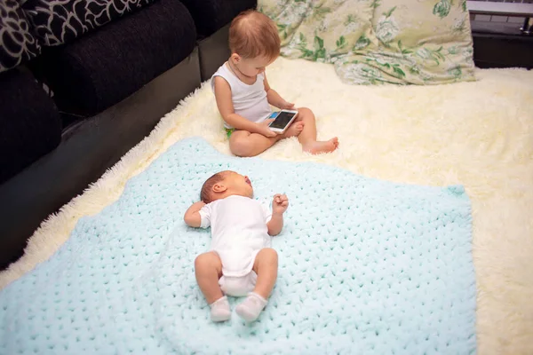 Um bebê recém-nascido está em um sofá, ao lado de um irmão de dois anos de idade joga um smartphone. Crianças e gadgets modernos — Fotografia de Stock