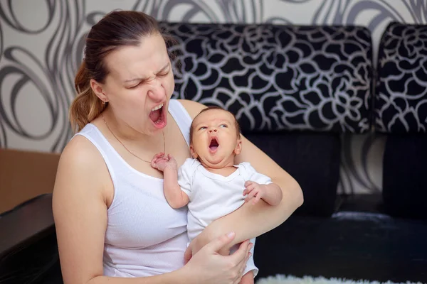 A young mother holds a newborn baby in her arms and yawns with him at the same time.