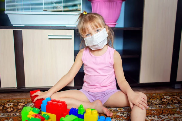 Little girl sits at home in quarantine during self-isolation in a medical mask and plays constructor