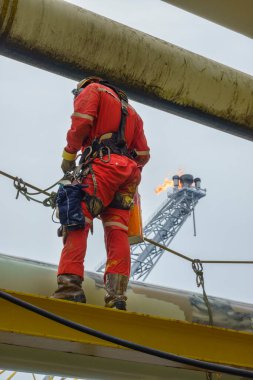 Working at height. A rear view of abseiler wearing Personal Protective Equipment (PPE) such as hard hat, harness, and coverall standing on the structure with background flare tip burning in the sky. clipart