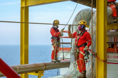 Working at height. A group of abseilers wearing red coverall and Personal Protective Equipment (PPE) such as hard hat, harness, hand protection and eye protection standing on the piepeline managing their rope access with background open sea. clipart