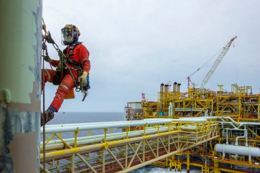 Yükseklerde çalışıyor. Solunum koruması, kasket, emniyet kemeri, el koruması, göz koruması ve arka plan yağ ve gaz platformu gibi kişisel koruyucu ekipman (PPE) giyen bir abseiler, boru hattında arka plan yağ ve gaz platformu ile resim faaliyetleri için asılı durumdalar.