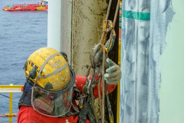 Yükseklerde çalışıyor. Solunum koruması, kasket, emniyet kemeri, el koruması, göz koruması ve arka planda açık deniz ile petrol ve gaz platformunun kenarında asılı duran kaplama gibi kişisel koruyucu ekipman (PPE) giyen bir abseiler.
