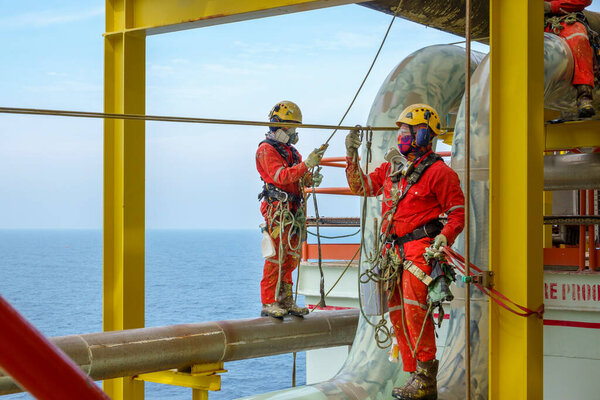 Working at height. A group of abseilers wearing red coverall and Personal Protective Equipment (PPE) such as hard hat, harness, hand protection and eye protection standing on the piepeline managing their rope access with background open sea.