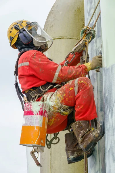 Working at height. An abseiler wearing Personal Protective Equipment (PPE) such as respiratory protection, hard hat, harness, hand protection, eye protection and coverall climbing at the edge of oil and gas platform for touch up painting on pipeline.