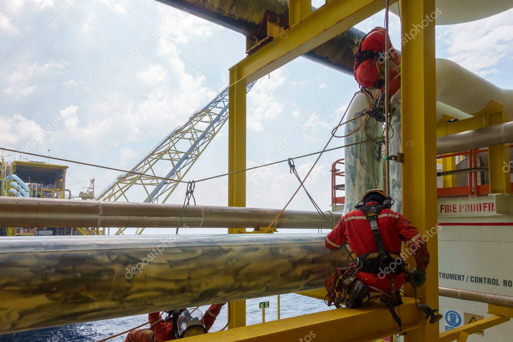 Working at height. An abseiler with red coverall and wearing Personal Protective Equipment (PPE) such as respiratory protection, hard hat, harness, hand protection, eye protection and coverall hanging at pipeline.