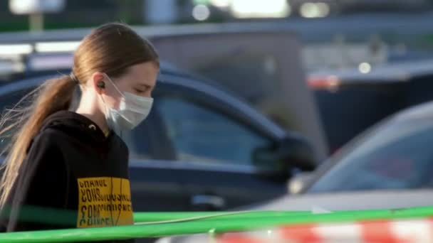 Gemaskerd jong meisje loopt geparkeerde auto coronavirus. Kind hoofdtelefoon gaat op straat covid-19 — Stockvideo