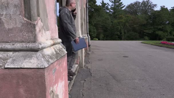 Чоловік з текою розмовляє на смартфоні і ходить біля старої будівлі — стокове відео