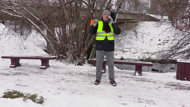 Školník s mluví o telefonu na chodníku poblíž lavičky lopata na sníh — Stock video