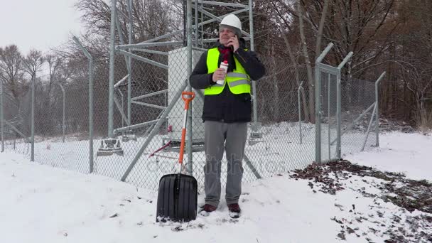 Trabalhador com telefone e garrafa térmica perto de pá de neve e cerca — Vídeo de Stock