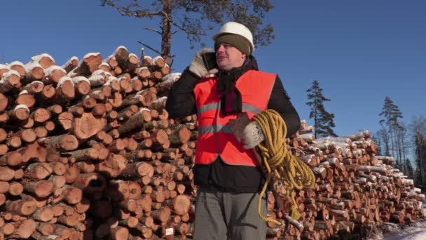 Lumberjack with rope and ax talking on phone near pile of logs in winter — Stock Video