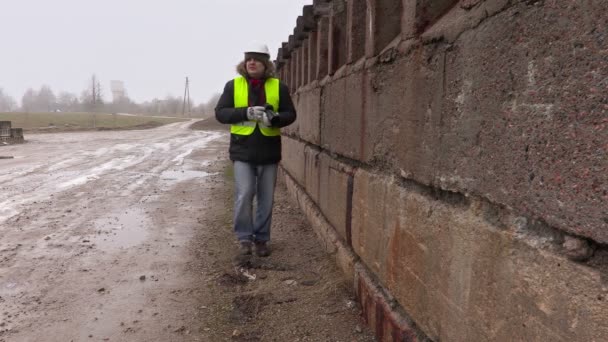 Инженер-строитель фотографирует на профессиональную фотокамеру и идет к бетонной стене — стоковое видео