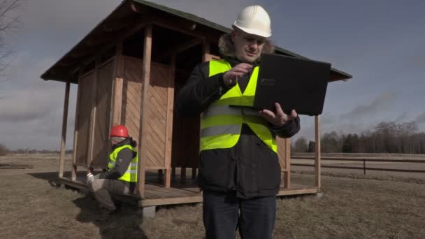 Працівники використовують ноутбук, ходьбу та каву біля дерев'яної будівлі — стокове відео