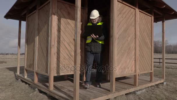 Construction engineer with laptop walking around wooden building — Stock Video