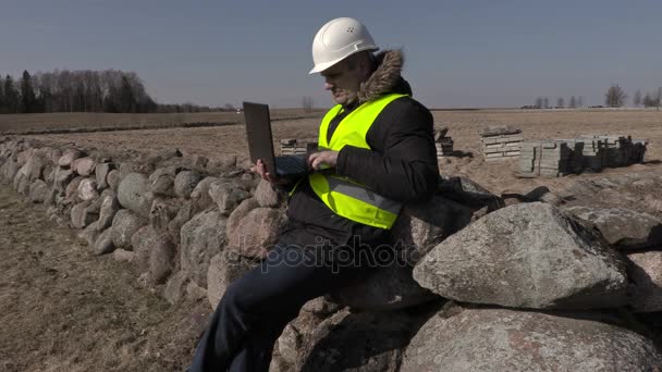 Инспектор здания сидит на каменном заборе и использует ноутбук — стоковое видео