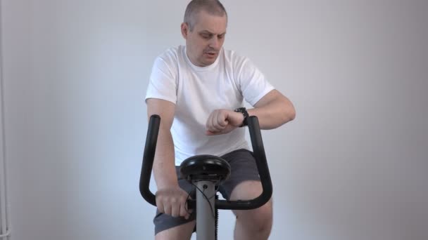 Hombre usando bicicleta estática y mira su reloj — Vídeo de stock